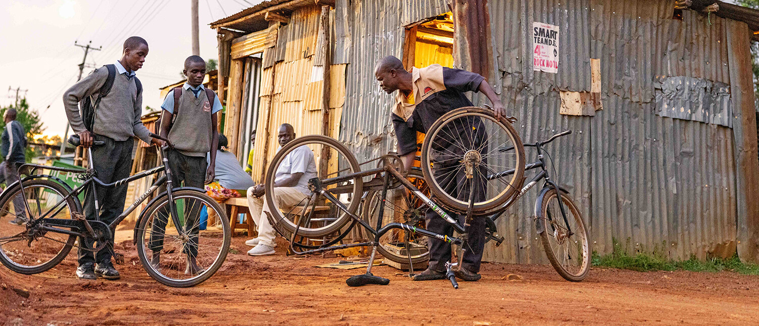 Several people gathered outside a small corrugated metal shop, with one man repairing a bicycle while two students in uniforms watch.