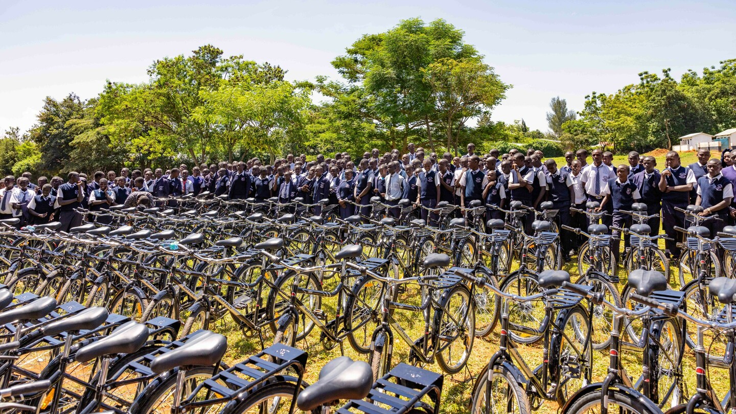 A large group of students in uniforms standing behind a row of stacked bicycles outdoors. Green trees and a clear sky are visible in the background.